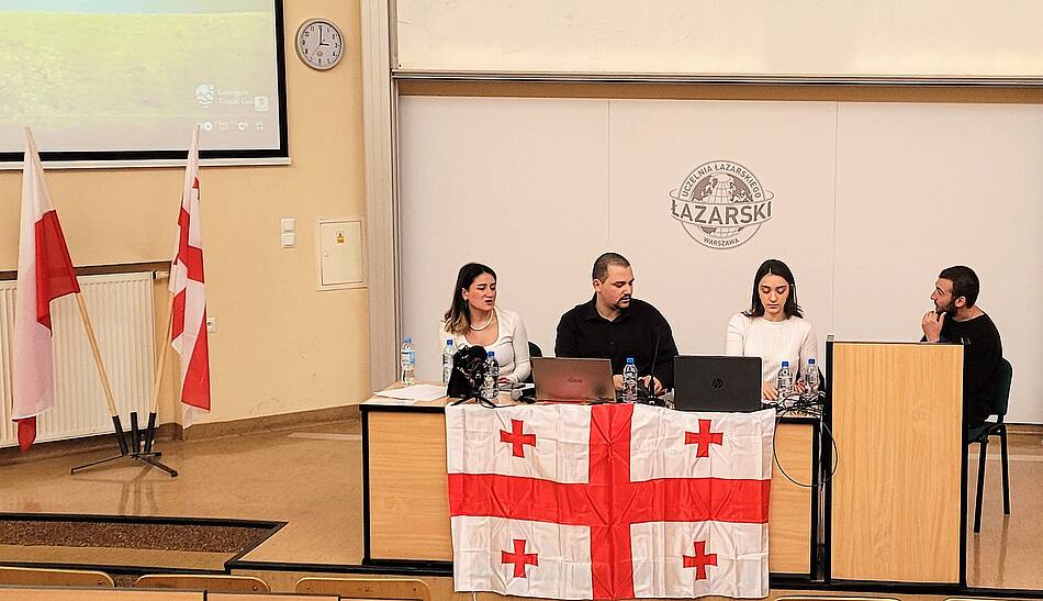 [Translate to Angielski:] Czterech studentów siedzi w audytorium na sali wykładowej za biurkiem. Na stole mają laptopa, powieszona jest też flaga Gruzji. Obok nich stoją dwie flagi, jedna polska, druga gruzińska.