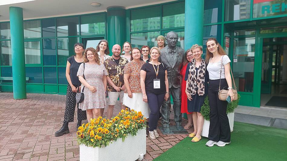 A group of smiling teachers stands in front of the Lazarski University building and poses for a photo at the monument to the founder of the Institution, Ryszard Lazarski