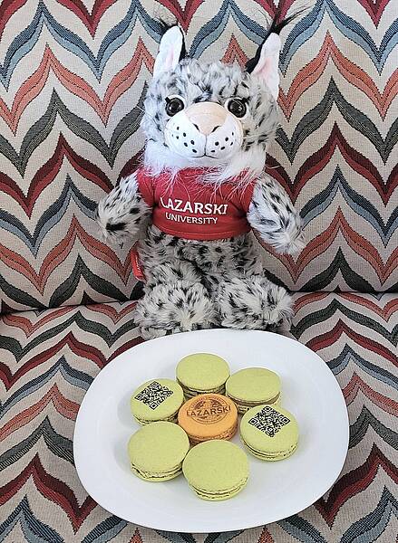 Lynx, a mascot of Lazarski University, is sitting on a chair. Next to his legs is a plate with French cookies - macarons.